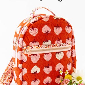May include: A quilted, orange backpack with a strawberry pattern. The backpack has a zippered front pocket, a top handle, and adjustable straps. The text "THE VIOLET BACKPACK" is at the top. The logo "KNOT + THREAD design" is at the bottom.