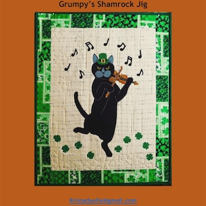 May include: A quilted wall hanging featuring a black cat playing a violin, wearing a green hat with a shamrock. The design includes musical notes and shamrocks on a cream background, framed by green and white patterned borders. The text reads "Trouble & Boo Designs" and "Grumpy's Shamrock Jig."