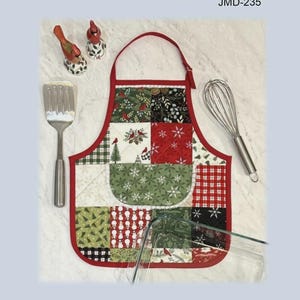 May include: Apron-shaped casserole hot pad with a patchwork design in red, green, and white. The apron has a red trim and a pocket. A spatula, whisk, and decorative birds are next to the apron. The text "Apron Casserole Hot Pad" is at the top.