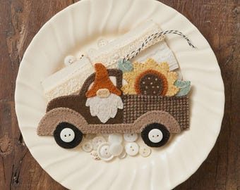 May include: A felt gnome truck decoration with a sunflower and lace accents. The truck is brown and tan, with a gnome wearing an orange hat. The text reads "Truckin' With My Gnomies Thru The Year {August}". Size: 3.5" x 6.25".