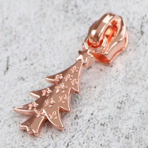 May include: Rose gold zipper pull with a Christmas tree design. The tree is decorated with stars.