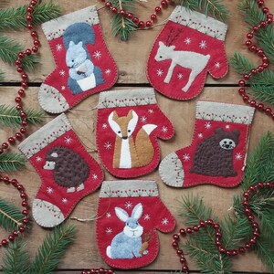 May include: A collection of six red felt Christmas ornaments in the shape of stockings and mittens. Each ornament features a different woodland animal: a squirrel, deer, fox, hedgehog, bear, and rabbit. The ornaments are decorated with white snowflakes.
