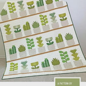 May include: A white quilt with a repeating pattern of green and gray potted plants. The quilt features the text "GREENHOUSE" and "Plant Sampler Quilt Pattern" with the designer's name, Elizabeth Hartman.