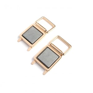 May include: Two gold-colored magnetic clasps with rectangular gray magnets. Each clasp has a rectangular frame with a hinged loop and two small tabs. The clasps are set against a white background.