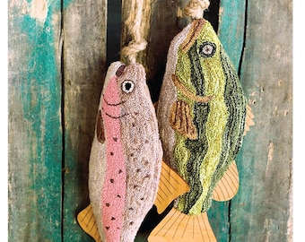 May include: Two plush fish ornaments, a trout and a bass, hanging from a rope with a red and white fishing bobber. The fish are made of a textured fabric and have embroidered details. The background is a weathered wooden surface.