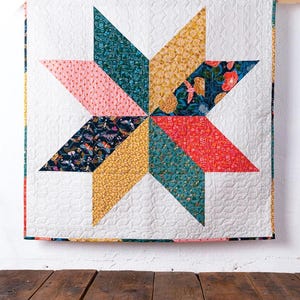 May include: A simple star quilt pattern with a white background and colorful fabric pieces in shades of pink, red, yellow, teal, and blue. The quilt is hanging on a white wall with a wooden floor below. The text "simple star quilt" is written below the quilt, and the text "a beginner-friendly oversized baby quilt" is written below that.