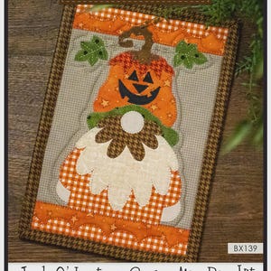 May include: A Jack-O'-Lantern Gnome mug rug kit. The kit features an orange pumpkin-shaped gnome with a smiling face, green leaves, and a white beard. The finished size is 7" x 10". The text "The Whole Country Caboodle" is at the top.