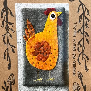 May include: A felt artwork of a yellow chicken with a red comb and a black eye. The artwork is framed in a gray square and has the text "Mrs. Hen" and "The Howells Nest #26" printed below it.