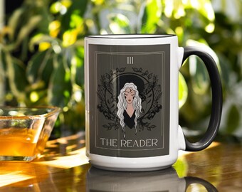 May include: White ceramic mug with a black handle. The mug features a graphic of a tarot card with a woman in a black dress and a white crown. The card is labeled "The Reader" and has a Roman numeral III in the top right corner.