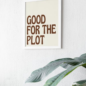 Pode incluir: Uma impressão emoldurada com a frase "GOOD FOR THE PLOT" em letras marrons sobre um fundo creme. A moldura branca está montada em uma parede branca texturizada, com folhas verdes de plantas em primeiro plano.