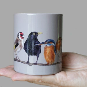 Puede incluir: Una taza de cerámica gris con una ilustración colorida de cuatro pájaros posados en una rama. Los pájaros son un jilguero, un mirlo, un martín pescador y un petirrojo.