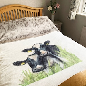 Puede incluir: Una manta blanca con una ilustración en blanco y negro de dos vacas en un campo de hierba verde.