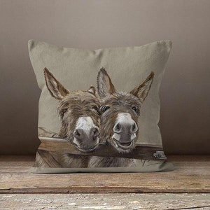 May include: A decorative throw pillow with a beige linen fabric cover featuring a hand-drawn illustration of two brown donkeys peeking over a wooden fence. The donkeys have a playful expression and are looking directly at the viewer.