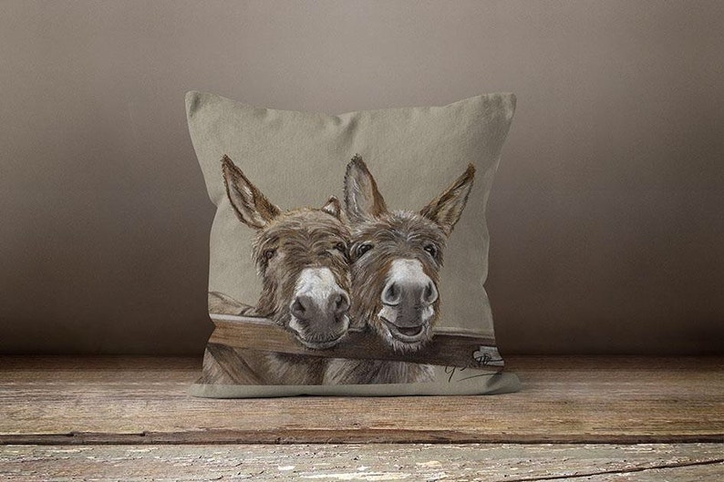 May include: A decorative throw pillow with a beige linen fabric cover featuring a hand-drawn illustration of two brown donkeys peeking over a wooden fence. The donkeys have a playful expression and are looking directly at the viewer.