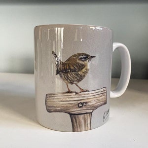 May include: A grey ceramic mug featuring a brown and white wren perched on a wooden post. The mug has a white handle and a brown and white illustration of the wren.