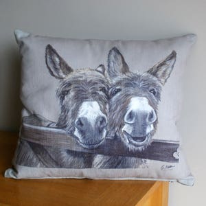 May include: A beige linen pillow with a printed image of two donkeys looking at the camera. The donkeys are brown and white with a wooden fence behind them.