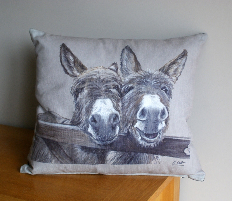 May include: A beige linen pillow with a printed image of two donkeys looking at the camera. The donkeys are brown and white with a wooden fence behind them.