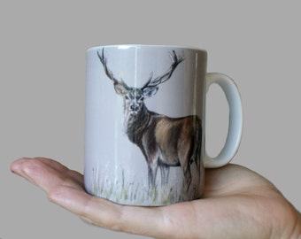 May include: A white ceramic mug featuring a brown and white illustration of a red deer with antlers. The deer is standing in a field of grass.