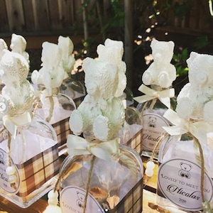 May include: Clear glass bottles with white teddy bear toppers, tied with cream-colored ribbons and twine. Each bottle has a plaid patterned label and a tag that reads "Lovely to Meet You" and a name. A decorative item for a baby shower or party.