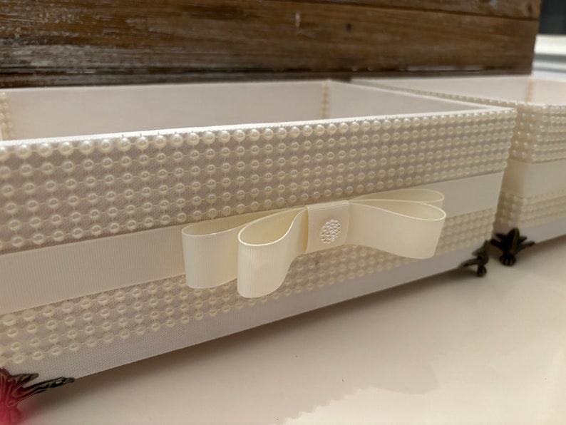 May include: Ivory-colored decorative boxes with pearl-like embellishments. A white ribbon with a bow adorns the front of the box. The boxes have a rectangular shape and are placed on a white surface. The boxes are ideal for storage.