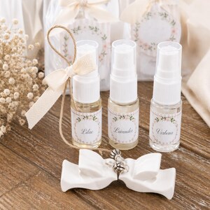 May include: Three small perfume bottles labeled "Lilac", "Lavender", and "Verbena" with floral labels. A white ceramic bow and a decorative bow with a tag are in the foreground. The background includes gift bags with floral tags.