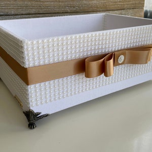 May include: A rectangular white box with pearl-like embellishments and a gold ribbon. The box has decorative metal feet and a gold bow with a crystal-like center. The interior is white.