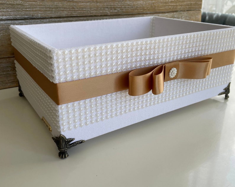 May include: A rectangular white box with pearl-like embellishments and a gold ribbon. The box has decorative metal feet and a gold bow with a crystal-like center. The interior is white.