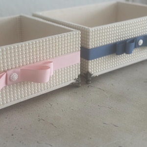 May include: Two rectangular decorative boxes. The boxes are covered in small, white, pearl-like beads. One box has a pink ribbon and bow, the other has a blue ribbon and bow. Both have a decorative button. The boxes are on a light-colored surface.
