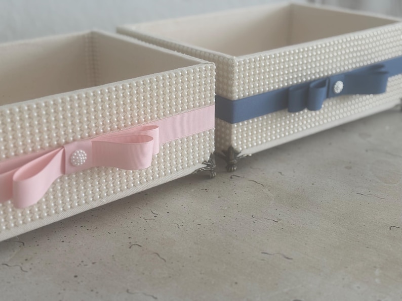 May include: Two rectangular decorative boxes. The boxes are covered in small, white, pearl-like beads. One box has a pink ribbon and bow, the other has a blue ribbon and bow. Both have a decorative button. The boxes are on a light-colored surface.