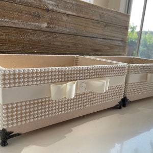 May include: Two rectangular cream-colored decorative boxes. The boxes are covered in small, evenly spaced, pearl-like beads. A cream-colored ribbon with a bow is wrapped around the center of each box. The boxes have ornate, dark-colored feet.
