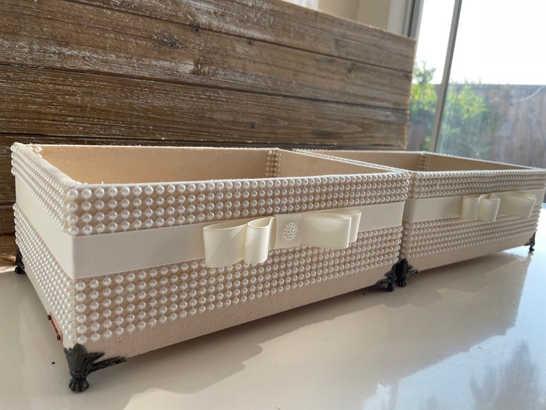May include: Two rectangular cream-colored decorative boxes. The boxes are covered in small, evenly spaced, pearl-like beads. A cream-colored ribbon with a bow is wrapped around the center of each box. The boxes have ornate, dark-colored feet.
