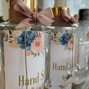 May include: Clear glass hand soap bottles with gold accents, floral labels, and pink bows. The labels feature blue and pink flowers with gold text that reads "Hand Soap". Each bottle has a gold chain with a crystal charm.