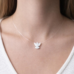 May include: A delicate white angel pendant necklace. The pendant is a small, detailed sculpture of an angel with outstretched wings. The necklace features a thin, nearly invisible chain, creating a floating effect. The angel pendant is centered on the chest.