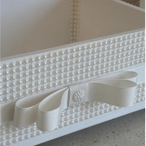 May include: A white rectangular box with a pearl-studded exterior. A white ribbon with a bow and a pearl-covered button adorns the front. The box is likely used for storage or decoration.