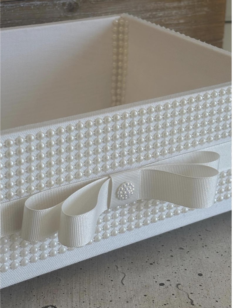 May include: A white rectangular box with a pearl-studded exterior. A white ribbon with a bow and a pearl-covered button adorns the front. The box is likely used for storage or decoration.