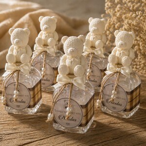 May include: Several clear glass bottles with teddy bear toppers, tied with cream-colored bows and twine. Each bottle has a decorative label with the text "Lovely to Meet You" and a name. The bottles are arranged on a wooden surface.
