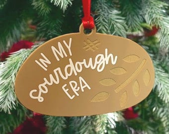 May include: A gold-colored oval Christmas ornament with the text "IN MY SOURDOUGH ERA" in white script. The ornament is hanging from a red ribbon against a backdrop of a Christmas tree with red accents.