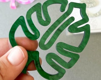 May include: A green, acrylic monstera leaf held in a hand, with a rainbow-colored leaf and a glittery leaf in the background. The glittery leaf is in a clear package with the text "be kind to yourself".