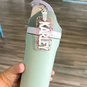 May include: A light green water bottle with a cactus pattern on the bottom. The bottle has a light purple handle and a clear tag that says "KARLEY" in white letters. The tag is attached with a silver clasp.
