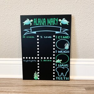 May include: A black chalkboard with a blue banner and green sea turtle illustrations. The chalkboard has white dots and text that reads "ALANA MARY", "I can", "I love", "I STAND", "I WEIGH", "Highlights", "I dislike", "I HAVE", and "TEETH".