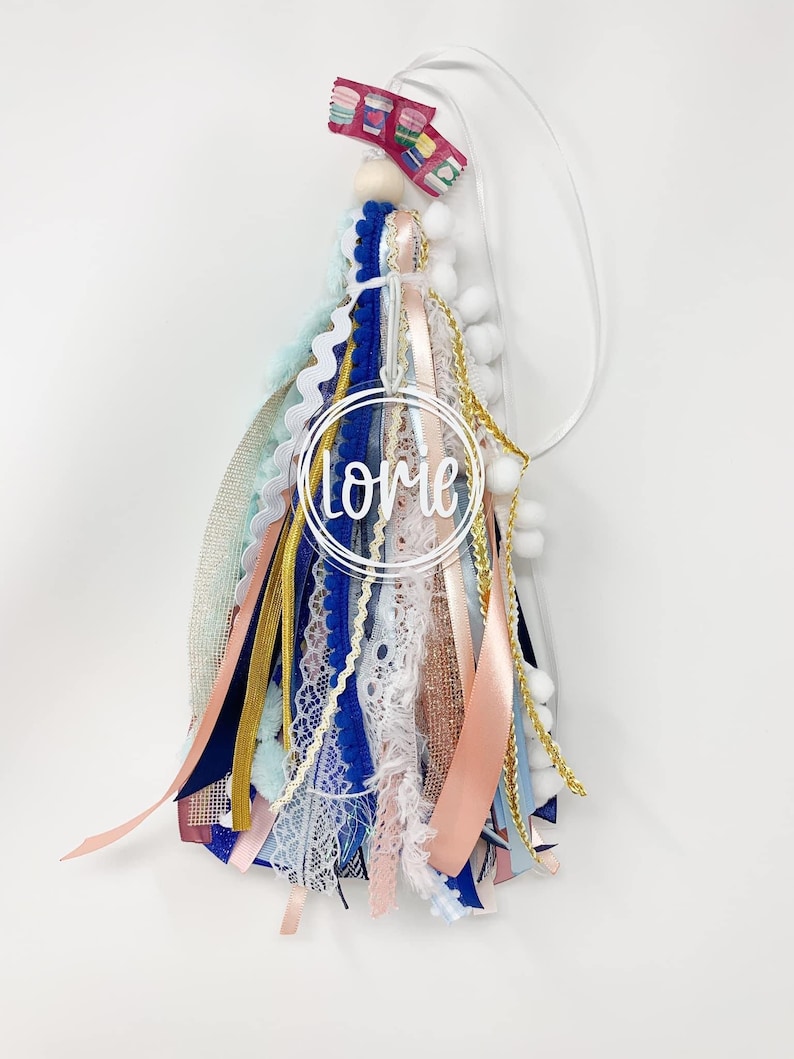 May include: A blue, pink, white, and gold tassel keychain with a clear circle charm that says "Lorie" in white lettering.