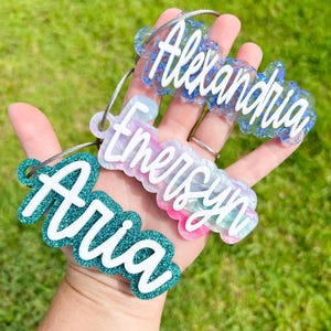 May include: Three personalized keychains with names "Aria", "Emersyn", and "Alexandria" written in white script on colorful resin backgrounds. The keychains are attached to silver metal rings.