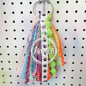 May include: A colorful key chain with a white circle that says "Angela" and black paw prints. The key chain has a variety of ribbons, including pink, blue, green, yellow, and orange.