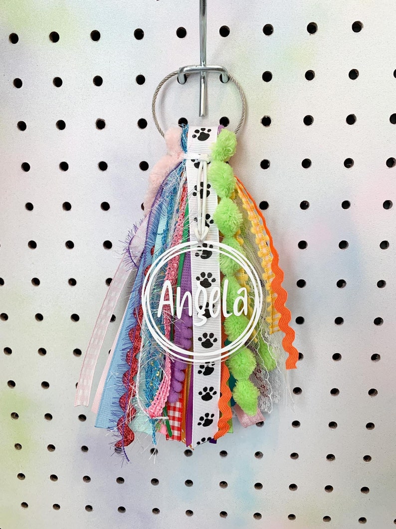 May include: A colorful key chain with a white circle that says "Angela" and black paw prints. The key chain has a variety of ribbons, including pink, blue, green, yellow, and orange.