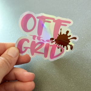 May include: A pink and iridescent sticker with the text "OFF the GRID" in a stylized font. A brown paint splatter is behind the text.