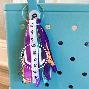 May include: A turquoise tote bag with a white circle charm with the letter "L" and paw prints. The charm is attached to a colorful tassel with ribbons and fringe.