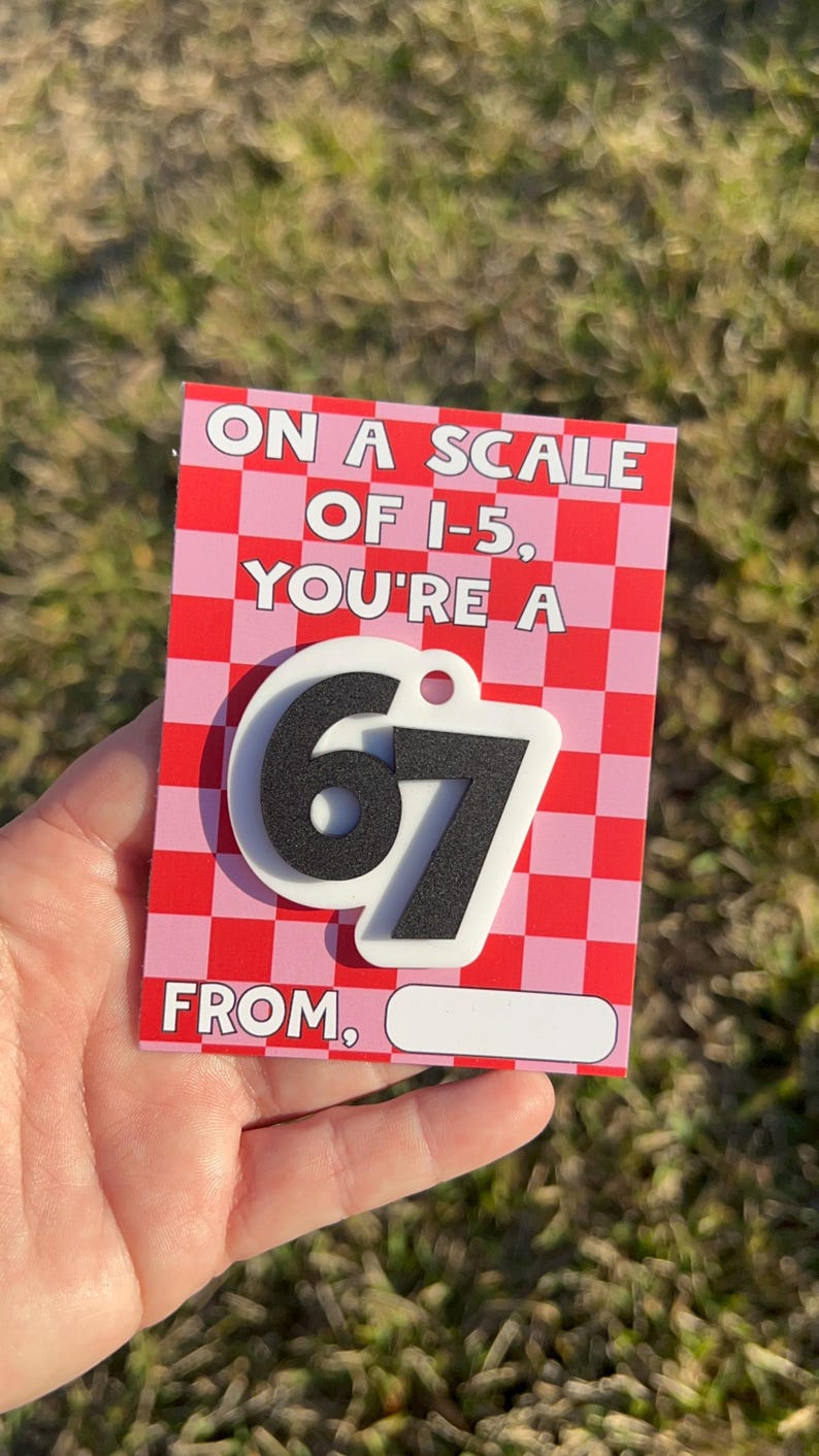 May include: A red and pink checkered card with the text "ON A SCALE OF 1-5, YOU'RE A" and a large "67" in black and white. The card also has the word "FROM" and a blank space for a name.
