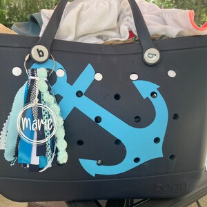 May include: A black tote bag with a blue anchor design and white polka dots. The bag has a white personalized tag that says "marie" and a blue and white tassel.