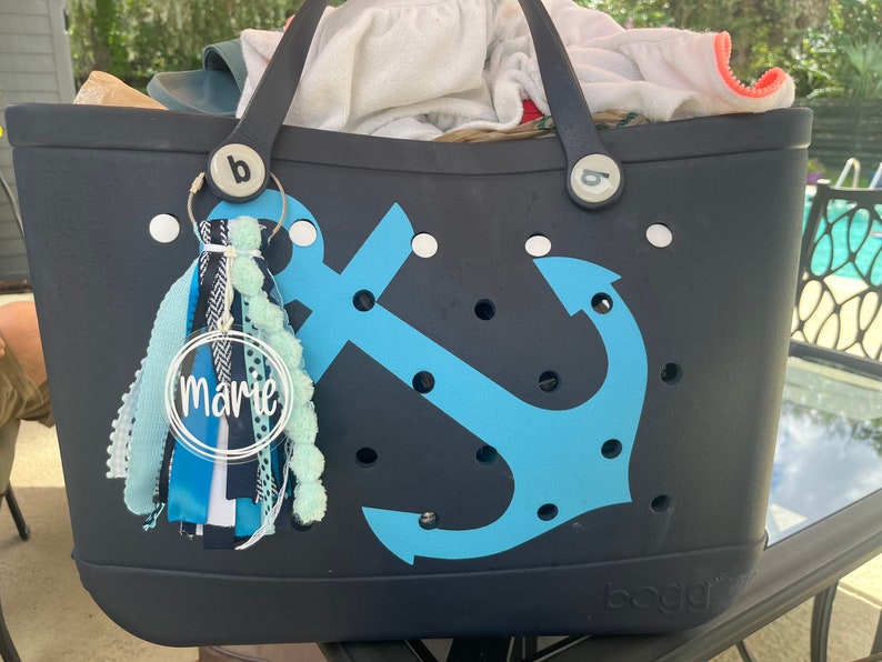 May include: A black tote bag with a blue anchor design and white polka dots. The bag has a white personalized tag that says "marie" and a blue and white tassel.