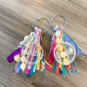 May include: Two clear acrylic keychains with colorful tassels. The keychains have the names "Hailey" and "Candace" written in white on them.